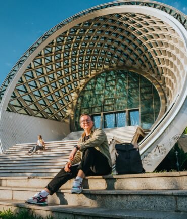 Purposeful young man sitting on stairs against modern building on sunny day dreaming about future,